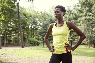 Smiling woman with hands on hip looking away while standing on field in park - CAVF01603