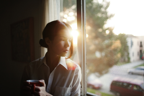 Frau schaut weg, während sie zu Hause am Fenster sitzt, lizenzfreies Stockfoto