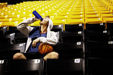Mann mit Basketball trinkt Wasser, während er auf einem Sitz im Stadion sitzt - CAVF01948