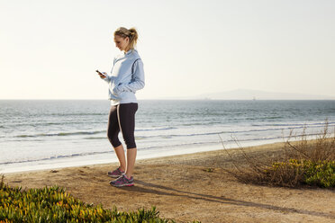 Seitenansicht einer Frau, die ihr Smartphone benutzt, während sie am Ufer des Strandes steht - CAVF01957