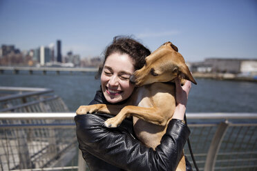 Happy woman embracing dog while standing on footbridge against sky - CAVF02009