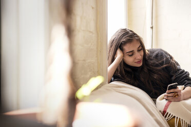 Woman with hand in hair using smart phone while leaning on sofa at home - CAVF02073