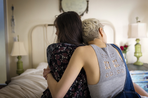 Rear view of friends embracing while sitting on bed at home stock photo