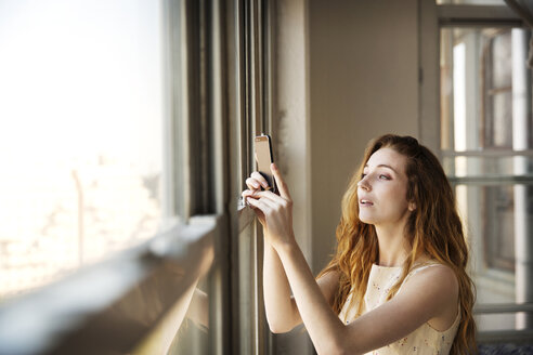 Woman photographing while standing by window at home - CAVF02103