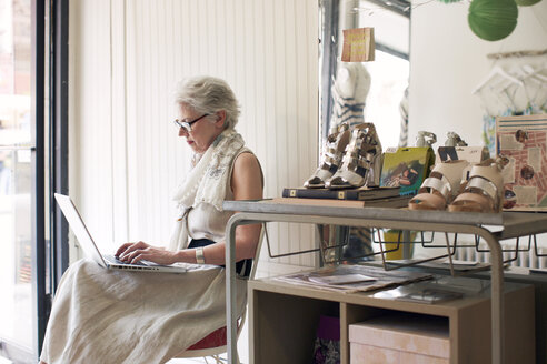 Seitenansicht einer älteren Frau, die einen Laptop benutzt und auf einem Stuhl in einem Geschäft sitzt - CAVF02176