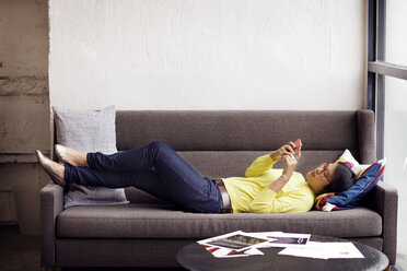 Seitenansicht einer Frau, die ein Smartphone benutzt, während sie auf dem Sofa im Büro liegt - CAVF02225