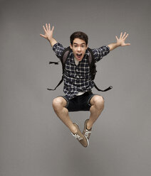 Portrait of cheerful man with backpack jumping against gray background - CAVF02336