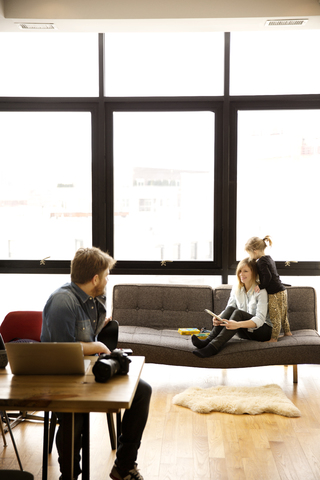 Frau schaut Mann an, während sie mit ihrer Tochter zu Hause auf dem Sofa sitzt, lizenzfreies Stockfoto