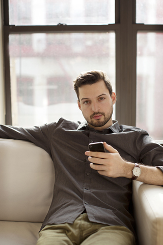 Porträt eines Mannes, der sein Smartphone hält, während er zu Hause auf dem Sofa sitzt, lizenzfreies Stockfoto