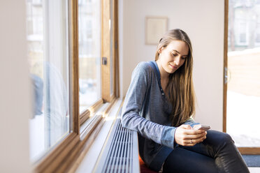Frau benutzt Smartphone, während sie zu Hause am Fenster sitzt - CAVF02540