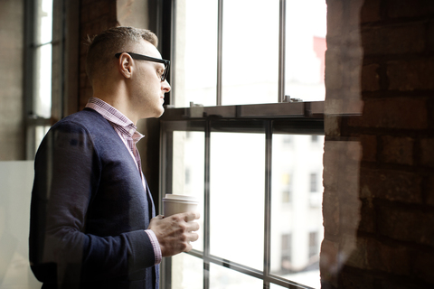 Geschäftsmann hält Einwegbecher, während er am Fenster im Büro steht, lizenzfreies Stockfoto