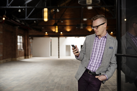 Geschäftsmann, der sein Smartphone benutzt, während er in einem leeren Lagerhaus vor der Tür steht, lizenzfreies Stockfoto