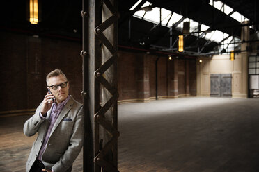 Businessman talking on smart phone while standing in empty warehouse - CAVF02638