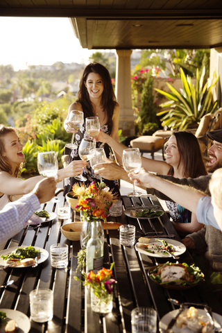 Glückliche Freunde stoßen mit Weingläsern am Esstisch auf der Veranda an, lizenzfreies Stockfoto