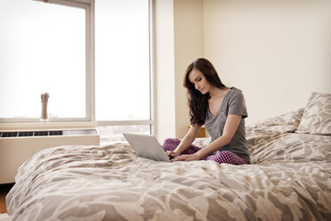 Seitenansicht einer Frau mit Laptop auf dem Bett in einem hell erleuchteten Schlafzimmer - CAVF02852