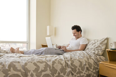 Seitenansicht eines glücklichen Mannes, der einen Laptop auf dem Bett zu Hause benutzt - CAVF02861