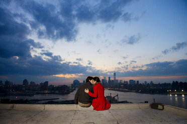 Rückansicht eines jungen Paares, das sich in der Abenddämmerung vor der Kulisse eines Flusses und einer Stadt küsst - CAVF02911