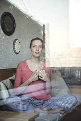Frau hält Kaffeetasse und sitzt auf Sofa durch Glasfenster gesehen - CAVF02913