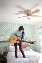 Glückliche Frau spielt Gitarre, während sie auf dem Bett zu Hause steht - CAVF02970