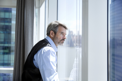 Nachdenklicher Geschäftsmann schaut durch das Fenster, während er in einem Hotelzimmer steht, lizenzfreies Stockfoto