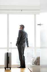 Thoughtful businessman standing with suitcase at window in hotel room - CAVF03012