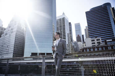 Businessman standing by railing against buildings in city on sunny day - CAVF03059