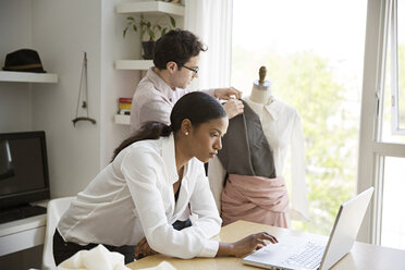 Eine Frau arbeitet am Laptop, während ihr Kollege im Studio ein Kleid an ein Modell näht - CAVF03103