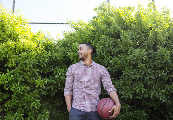 Happy young man holding basketball while standing against plants - CAVF03122