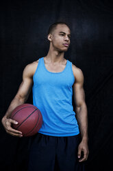 Selbstbewusster Sportler, der einen Basketball hält und gegen eine schwarze Wand schaut - CAVF03132