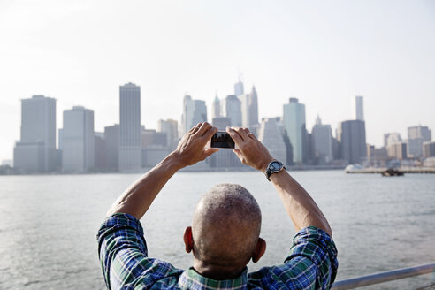 Rückansicht eines älteren Mannes, der die Stadt am East River mit seinem Smartphone fotografiert - CAVF03139