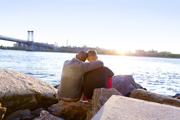 Rückansicht eines Mannes, der mit seinem Freund auf einem Felsen am East River bei Sonnenuntergang sitzt - CAVF03164
