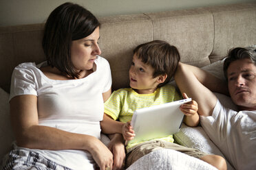 Boy using tablet computer while lying with parents on bed at home - CAVF03228