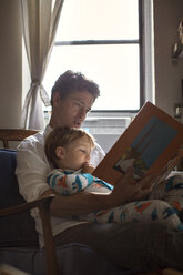 Vater liest seinem Sohn ein Bilderbuch vor, während er auf einem Stuhl am Fenster sitzt - CAVF03257