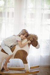 Portrait of smiling girl sitting on rocking horse against curtains at home - CAVF03268