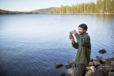 Mann, der am Seeufer stehend mit seinem Mobiltelefon fotografiert - CAVF03399