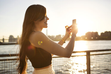 Glückliche Frau beim Fotografieren mit Handy am Fluss bei Sonnenuntergang - CAVF03762