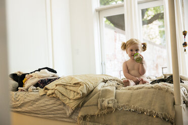 Portrait of baby girl holding bottle while sitting on bed at home - CAVF03841