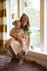 Mutter, die ihrer Tochter beim Anziehen der Schuhe hilft, während sie zu Hause am Fenster sitzt - CAVF03849