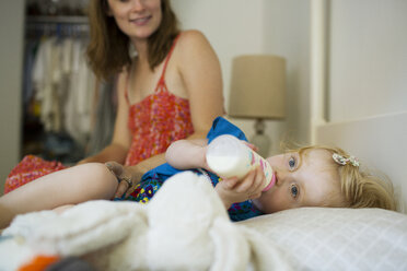 Baby girl drinking milk while mother sitting by on bed at home - CAVF03870