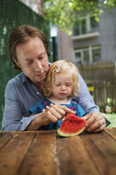 Father showing watermelon to daughter while sitting at table in backyard - CAVF03877