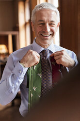 Smiling businessman trying on ties in mirror at menswear shop - CAIF08586