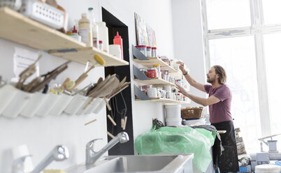 Mann stöbert im Töpferatelier in einem Regal mit Kunstzubehör - CAIF08633