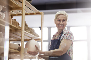 Lächelnde ältere Frau, die eine Töpfervase auf ein Regal im Atelier stellt - CAIF08646