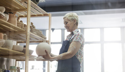 Ältere Frau stellt Töpfervase auf Regal in Atelier - CAIF08658