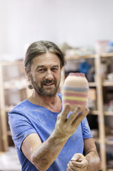 Porträt lächelnder reifer Mann mit bemalter Keramikvase im Atelier - CAIF08691