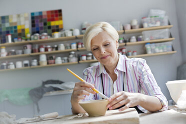 Smiling mature woman painting pottery bowl in studio - CAIF08694