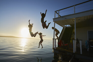 Junge erwachsene Freunde springen von einem Hausboot in den Sonnenuntergang im Meer - CAIF08786