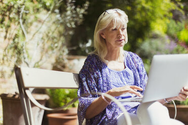 Ältere Frau mit Laptop auf einer sonnigen Gartenbank - CAIF08846