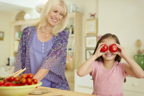 Porträt einer verspielten Enkelin, die sich in der Küche mit Tomaten die Augen zuhält - CAIF08851