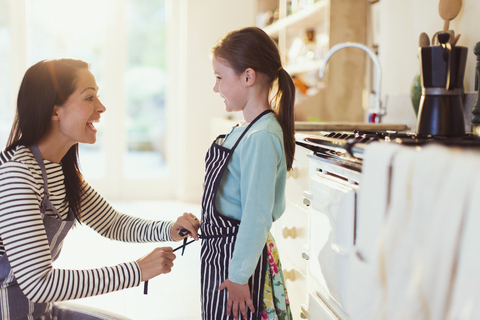 Mother tying apron on daughter in kitchen stock photo